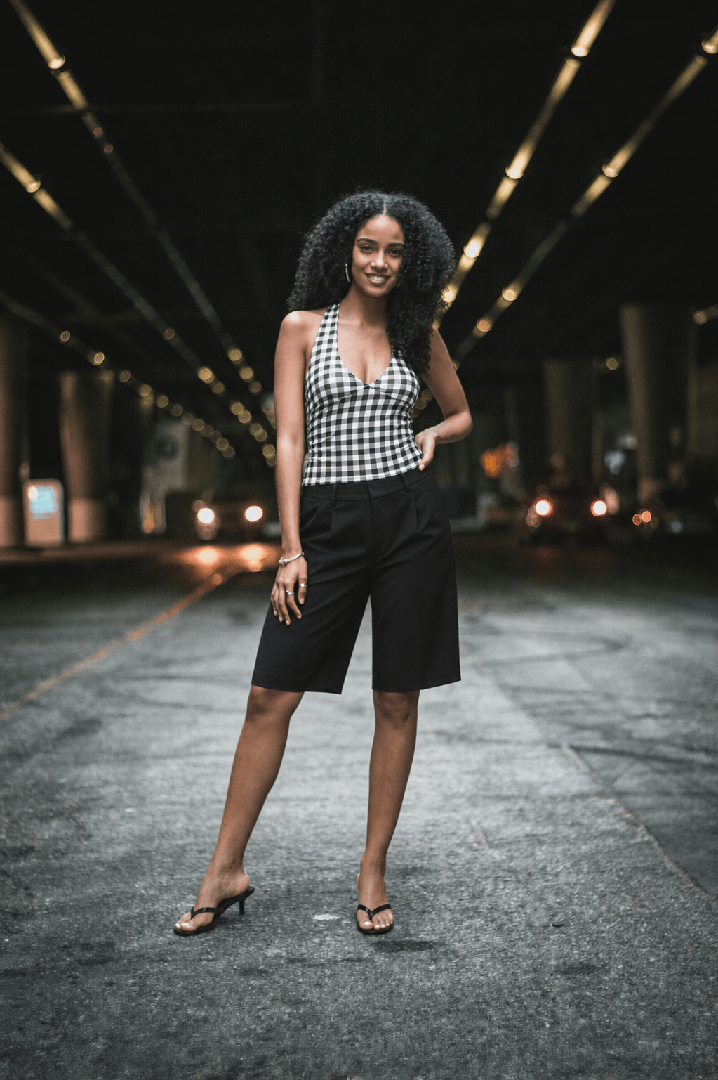 After: the same supplied real woman model in the unchanged photo, wearing the reference black-and-white gingham halterneck top, black tailored Bermuda shorts, and black kitten-heel mules